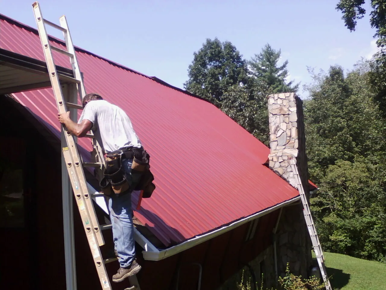 Roofer on a ladder beside a red metal roof and stone chimney — Soffit & Fascia Repair by Roofing Brothers in Stormville